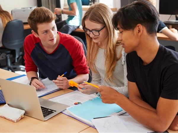 Drei junge Menschen arbeiten gemeinsam mit Laptop, Unterlagen und Stiften an einem Tisch.
