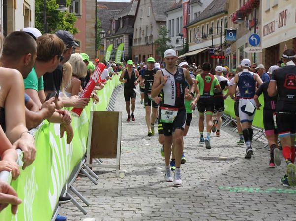 Teilnehmende eines Lauf- oder Triathlonwettbewerbs bewegen sich durch eine Innenstadt, während Zuschauer am Streckenrand stehen.