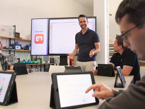 Lehrkraft präsentiert im Klassenzimmer, während Schülerinnen und Schüler mit Tablets arbeiten.