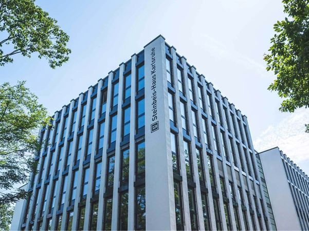 Modernes Bürogebäude mit Glasfassade, umgeben von Bäumen, mit Schriftzug an der Gebäudeecke.