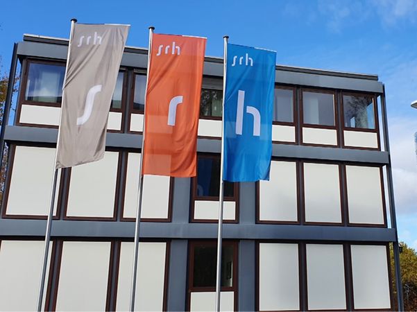 Gebäude der SRH-Gruppe mit farbigen Unternehmensflaggen vor blauem Himmel.