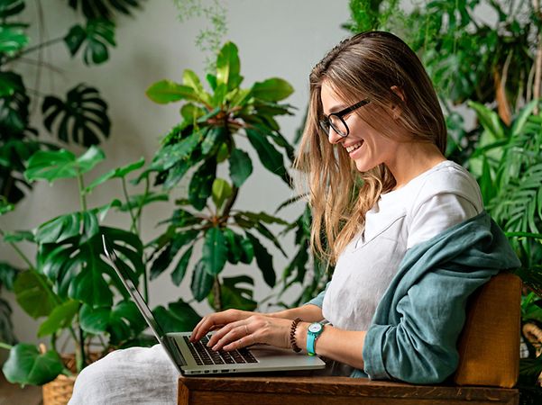 Person sitzt in einem Sessel und arbeitet mit einem Laptop, umgeben von grünen Zimmerpflanzen