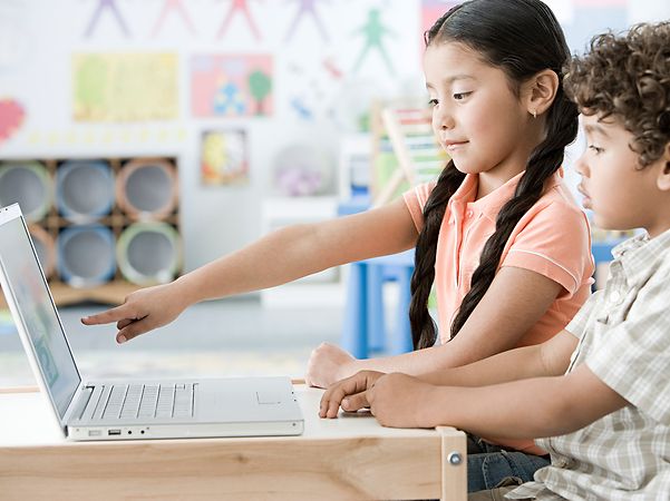 Zwei Kinder arbeiten im Klassenzimmer gemeinsam an einem Laptop, ein Mädchen zeigt auf den Bildschirm.