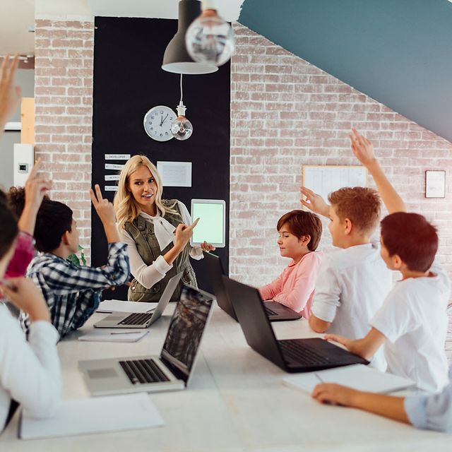 Lehrerin zeigt im Klassenzimmer ein Tablet, während mehrere Kinder mit Laptops sitzen und sich melden