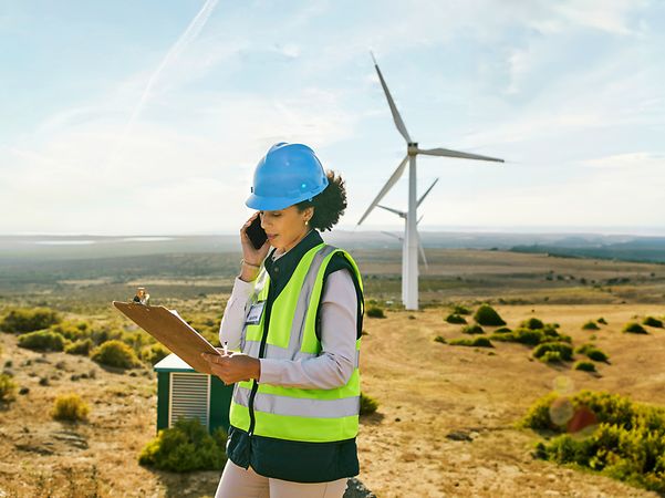 Frau mit Schutzhelm und Warnweste telefoniert mit Notizblock in einer Landschaft mit Windrädern.