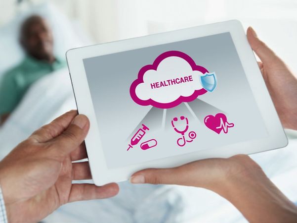 Zwei Personen halten ein Tablet mit digitalen Healthcare-Symbolen vor einem Krankenhausbett.