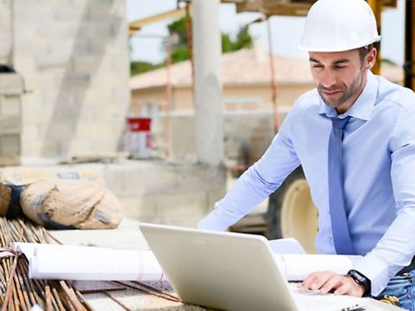Person mit Schutzhelm arbeitet auf einer Baustelle am Laptop neben Bauplänen.